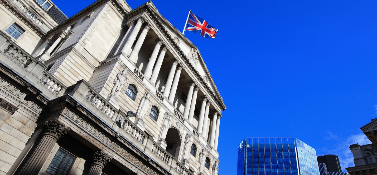 Image of the Bank of England building with a Union Jack flag flying on top, set against a bright blue sky, with modern glass skyscrapers in the background.
